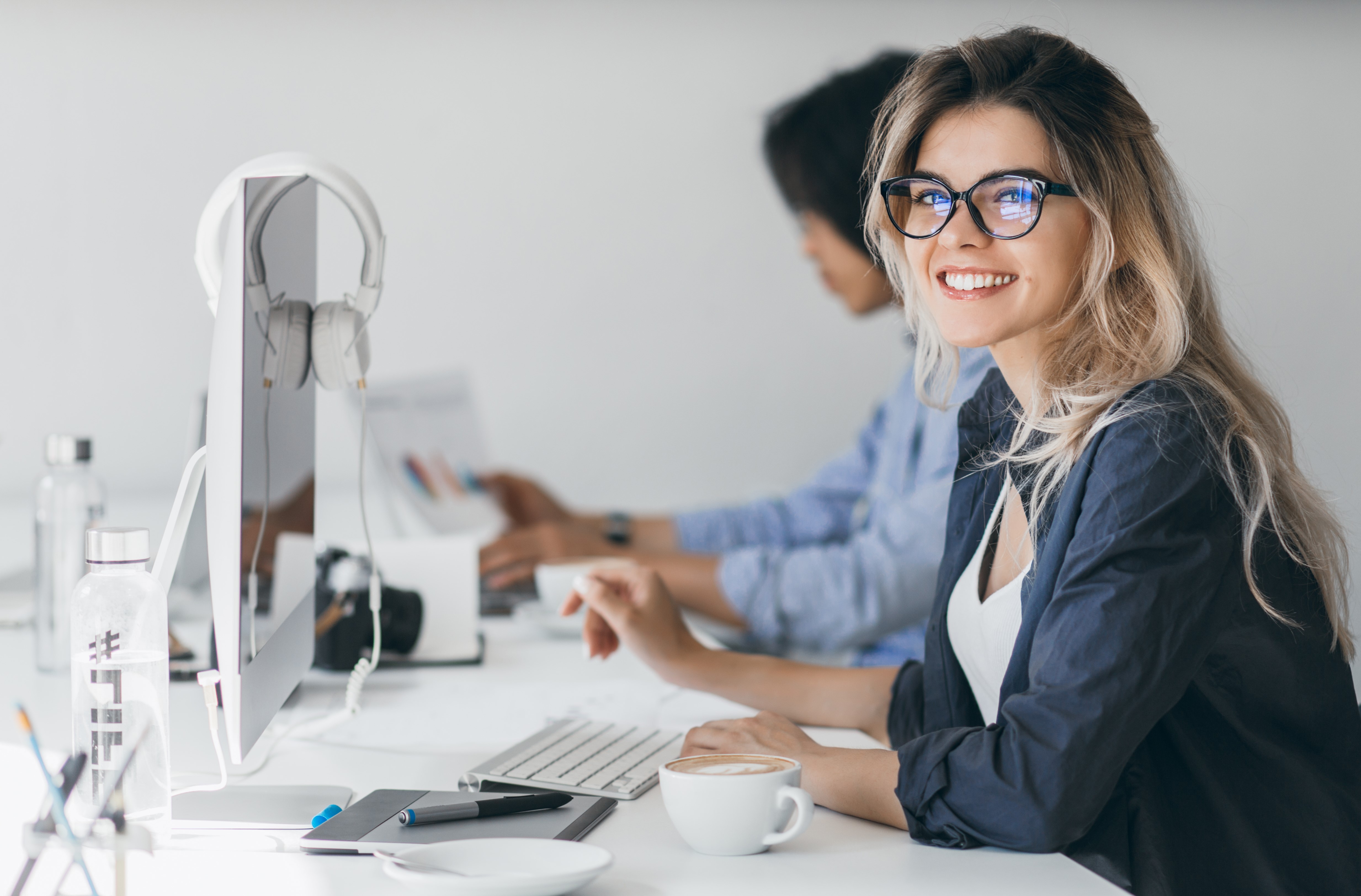 Attractive laughing freelancer girl posing with cup of coffee at her workplace. Chinese student in blue shirt works with document in campus with blonde friend in glasses.
